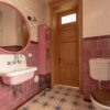 Interior of clean light restroom with retro sink beside big round mirror and decorated cabinet on tall wall with white and pink tile next to toilet bowl with brown lid in home