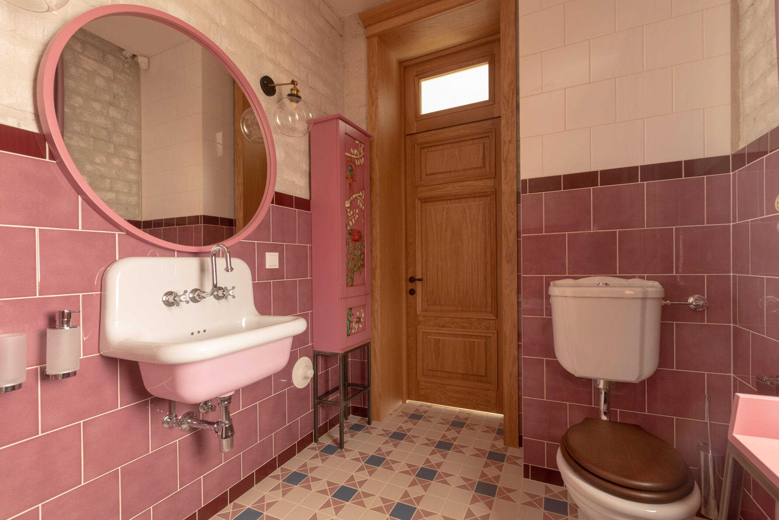 Interior of clean light restroom with retro sink beside big round mirror and decorated cabinet on tall wall with white and pink tile next to toilet bowl with brown lid in home