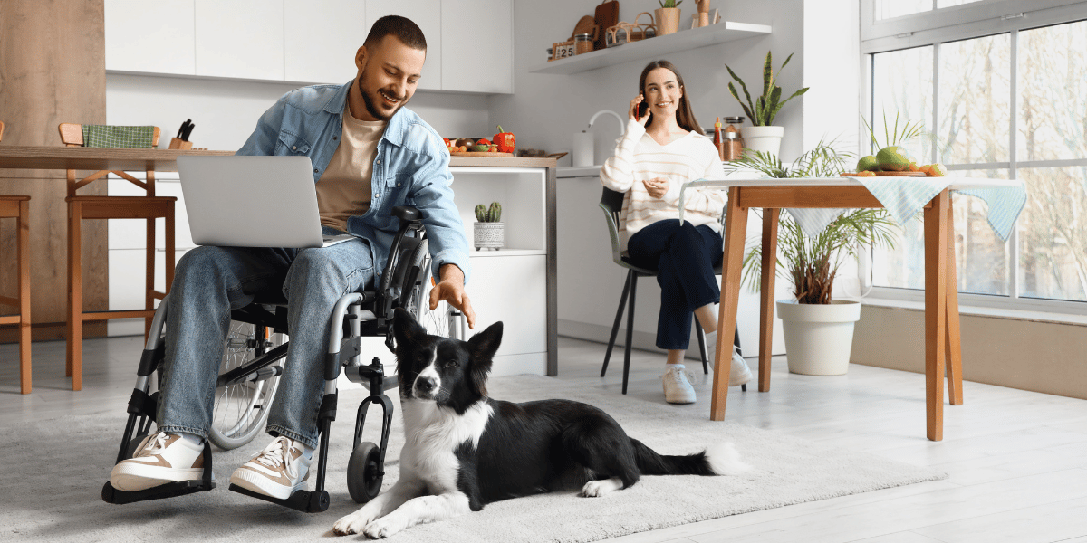 Handicap-Accessible Home Improvements_ When to Call in Expert Contractors - featured image man with laptop in wheelchair petting dog - woman on phone in background in a kitchen