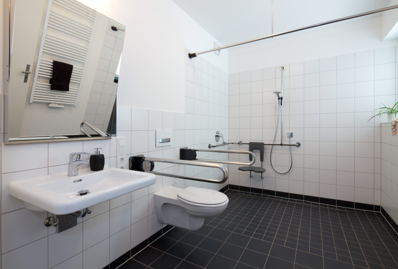 barrier-free bathroom renovations bowling green ky - a white and black tile bathroom showing a sink with mirror, a toilet with grab rails on either side, and a shower with a seat and grab bars